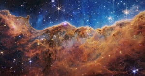 A colorful image of a star-forming region in space shows bright stars scattered across a blue and black sky above glowing, orange-brown clouds of gas and dust, resembling a mountainous landscape.
