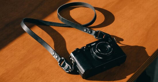 A black camera with a leather strap lies on a wooden surface, partially illuminated by sunlight, casting soft shadows.