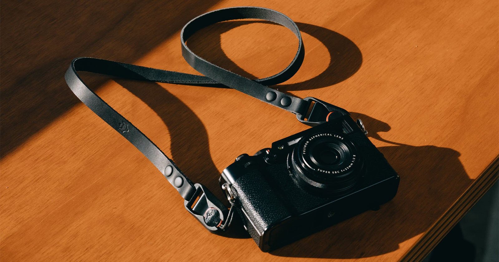 A black camera with a leather strap lies on a wooden surface, partially illuminated by sunlight, casting soft shadows.
