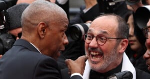 A man in a suit gestures animatedly toward another man with glasses and a white scarf, who is smiling widely and appears excited. Photographers and cameras surround them in the background.