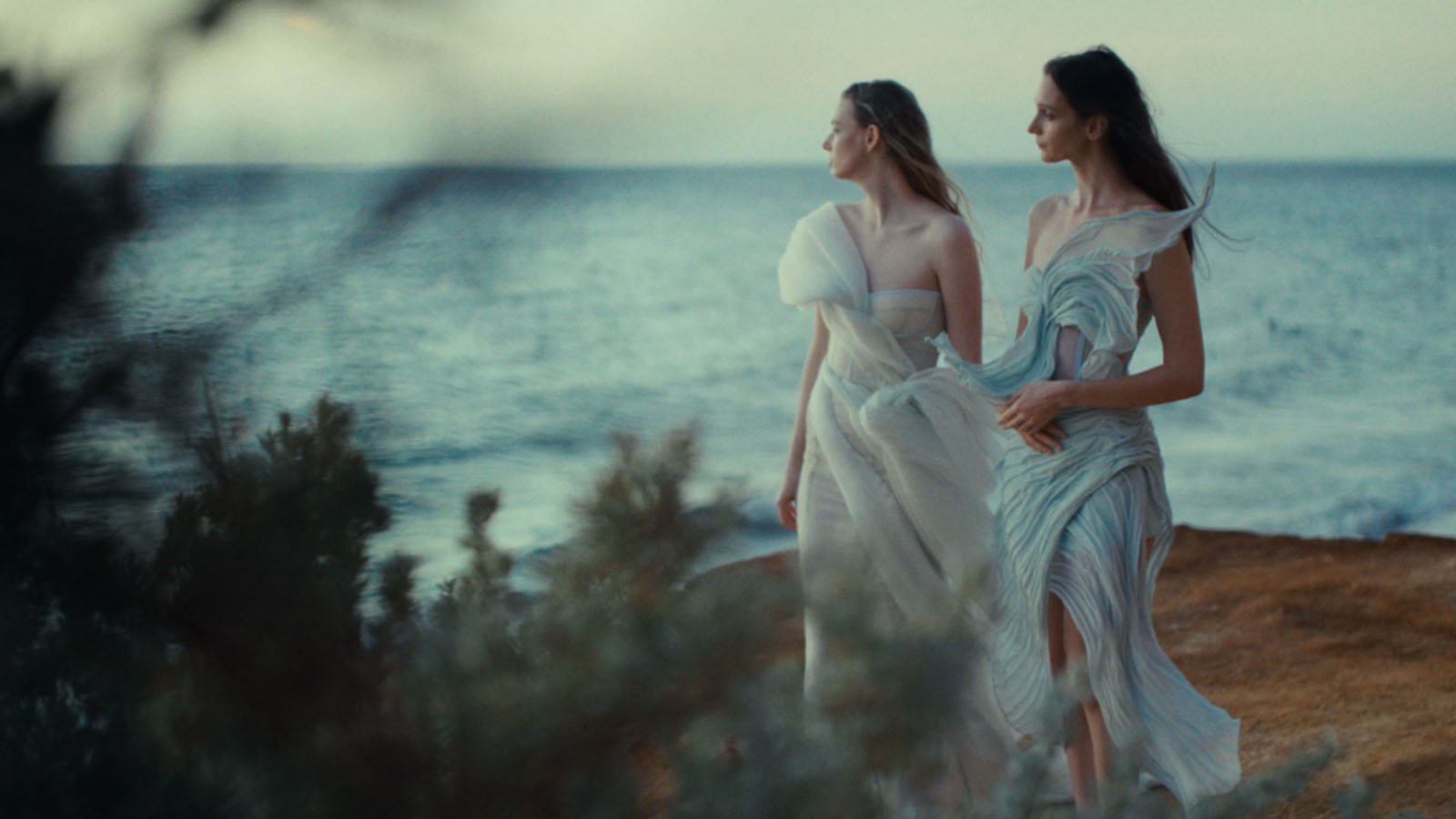 Two women in flowing, pastel-colored dresses stand on a rocky shoreline, looking out over the ocean at sunset. Soft greenery and blurred plants frame the foreground, creating a dreamy, ethereal atmosphere.