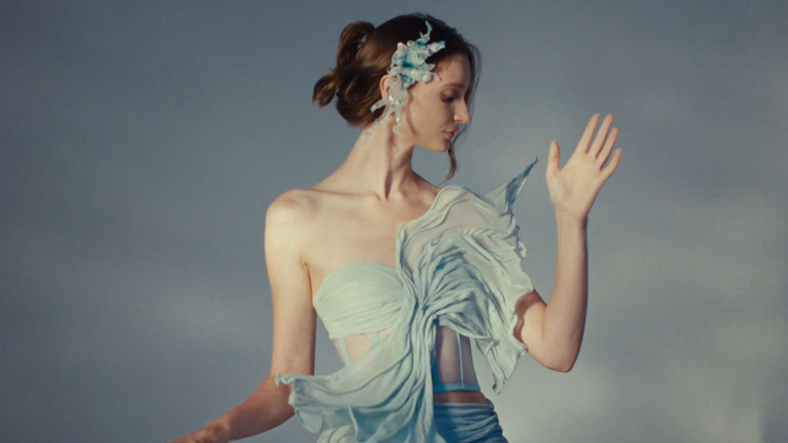 A woman in a flowing, light blue, off-shoulder dress poses gracefully against a soft gray background. She wears a delicate floral hair accessory and raises one hand, looking down with an elegant expression.