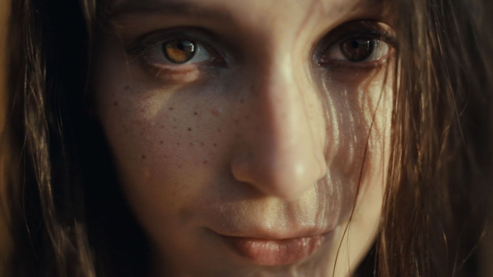 A close-up of a young person’s face with brown eyes, freckles, and light skin. Strands of dark brown hair fall across their face, and they have a neutral expression. Soft natural light illuminates their features.