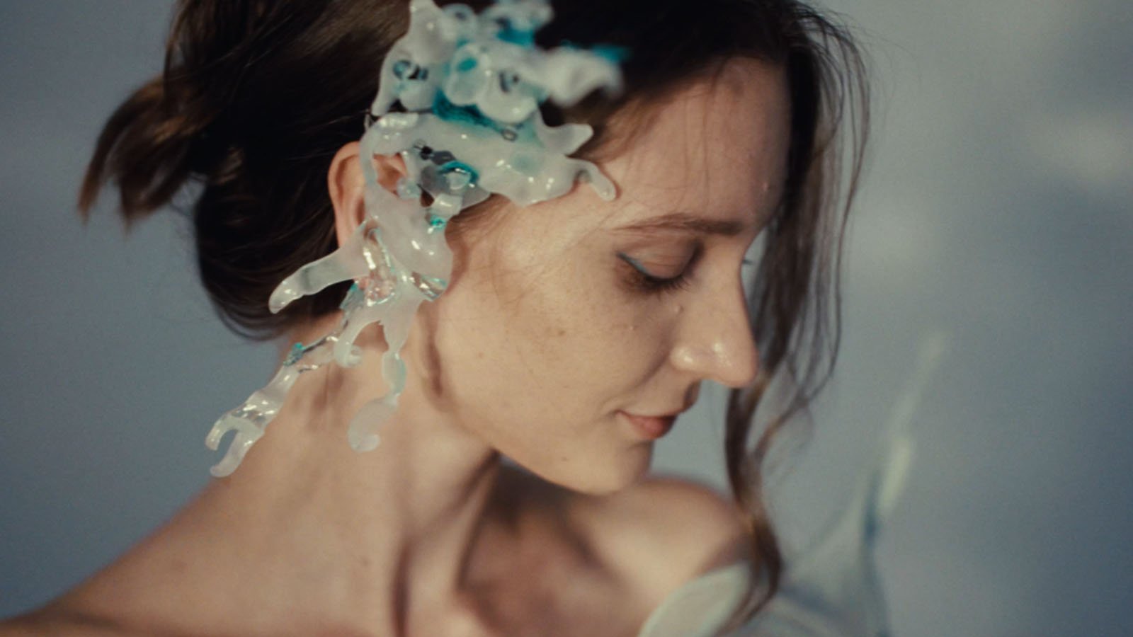 A woman with closed eyes and a gentle expression has translucent, blue and white sculptural art pieces attached to the side of her face and ear. Soft lighting highlights her serene profile.