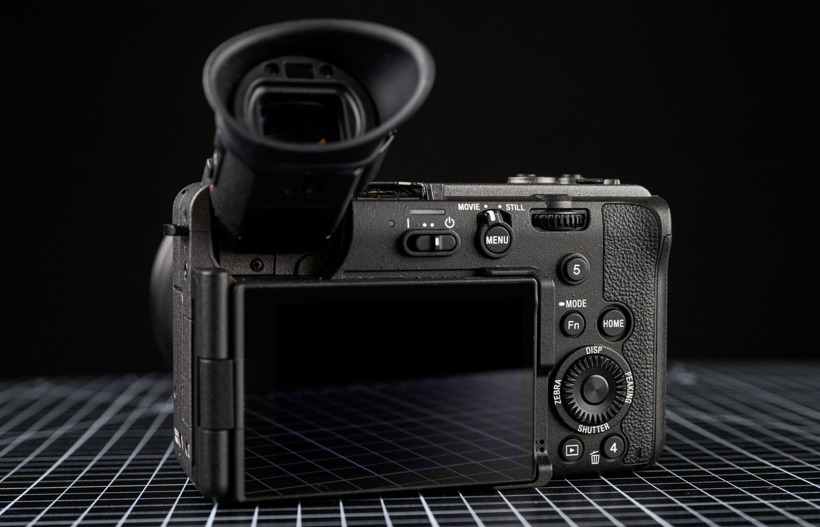 A digital camera viewed from the back, showing its buttons, dials, and a flip-out LCD screen, placed on a grid-patterned surface with a black background.