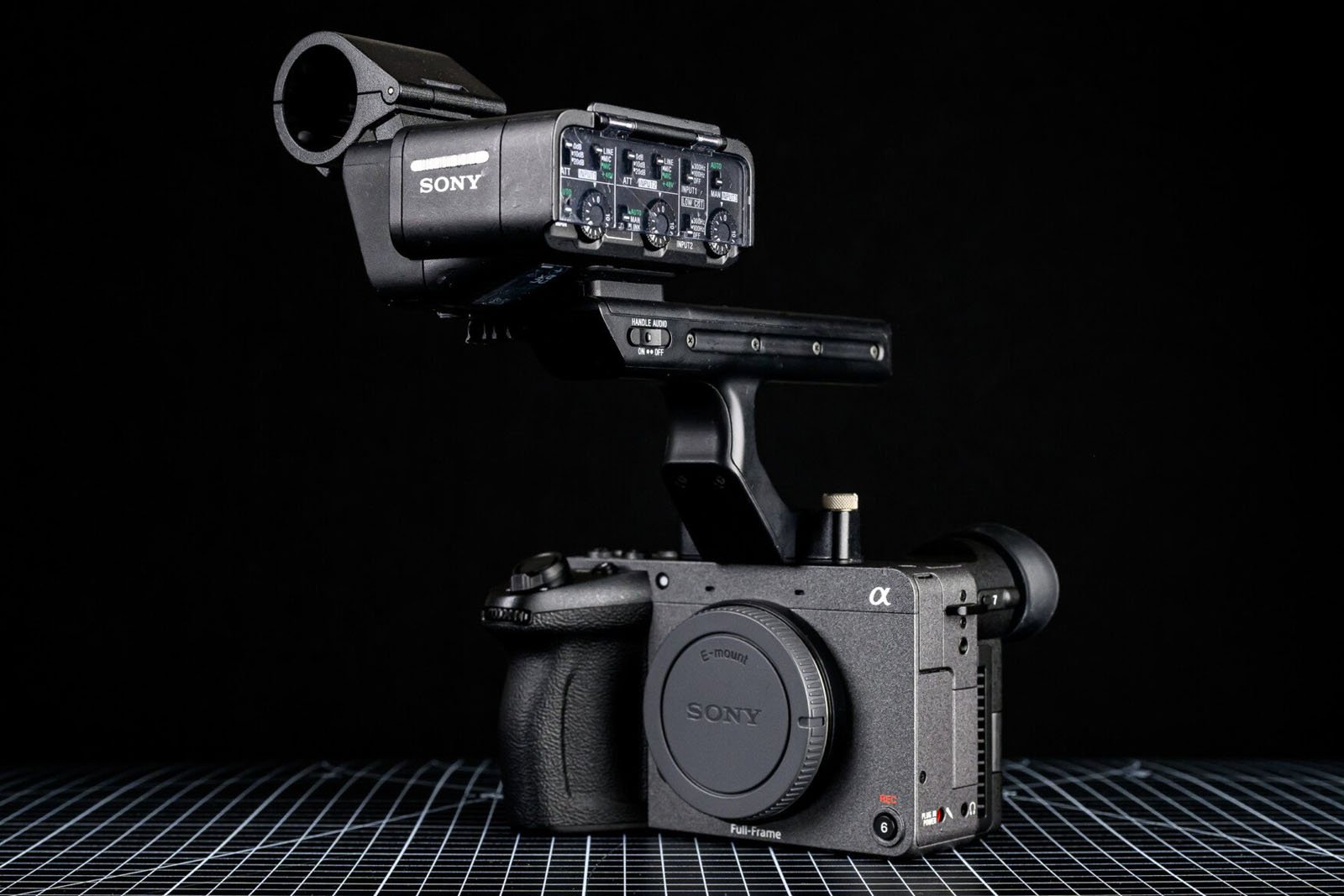 A Sony Alpha camera with an external microphone and audio interface attached, displayed against a black background on a grid-patterned surface.