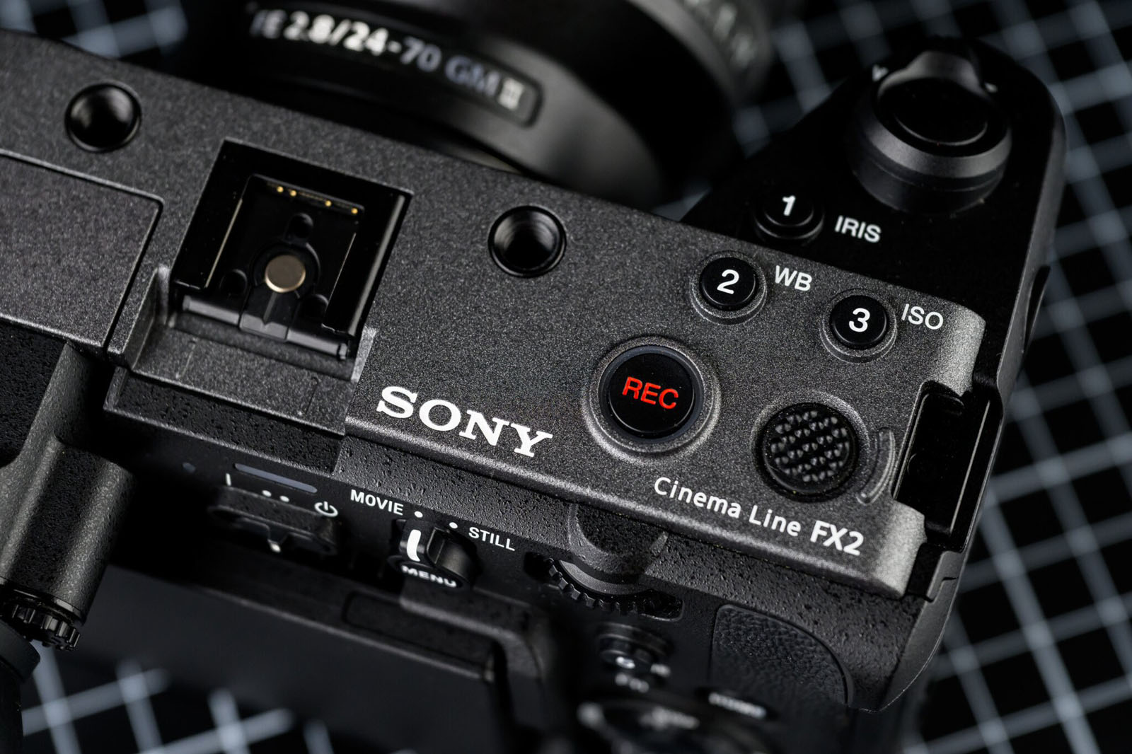 Close-up of the top controls of a Sony Cinema Line FX2 camera, showing the REC button, dials for white balance and ISO, and part of the lens, with a grid background.