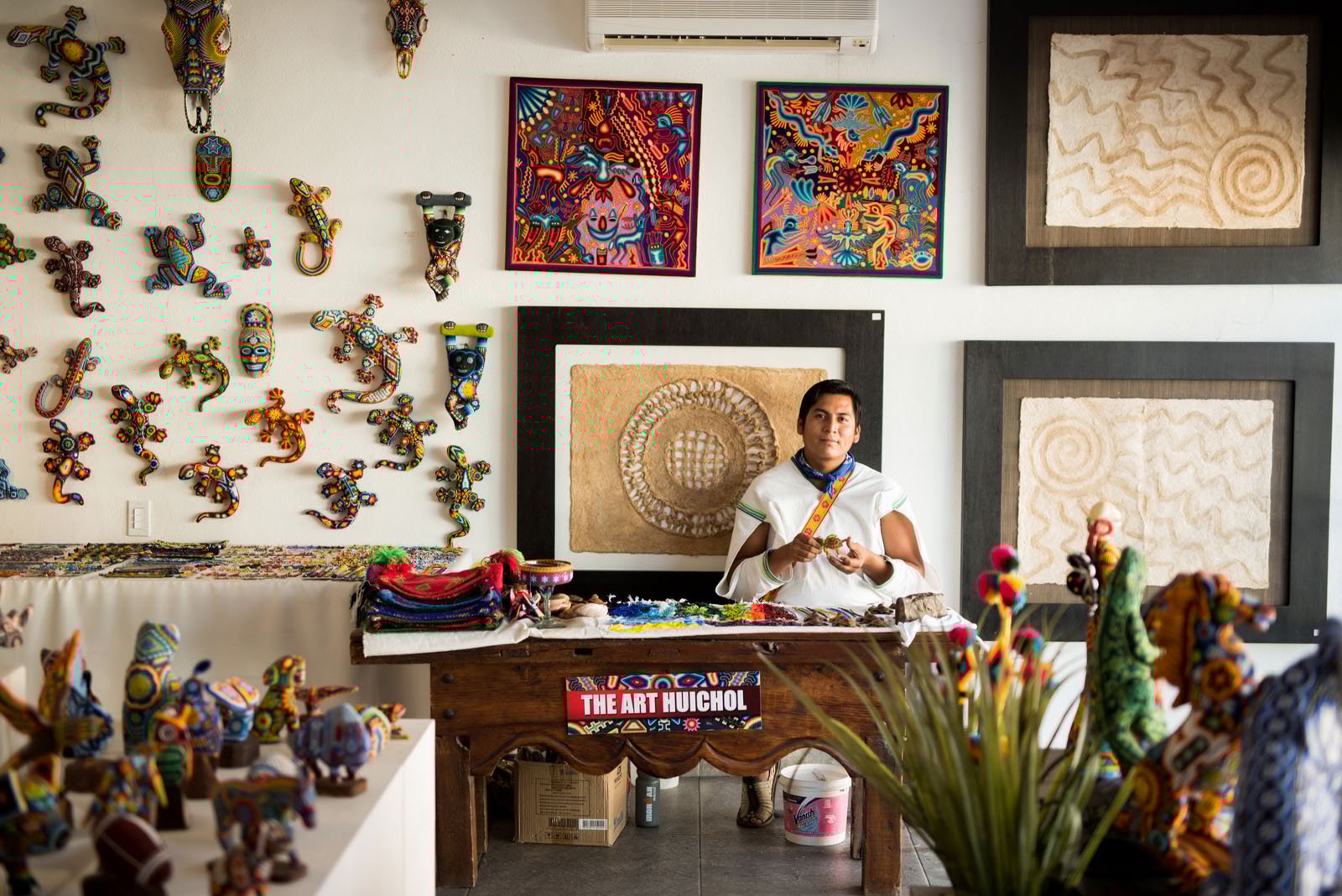 A woman in traditional clothing sits at a table displaying colorful Huichol art. The walls behind her are covered with vibrant beadwork, masks, and embroidered pieces, creating a lively, artistic atmosphere.