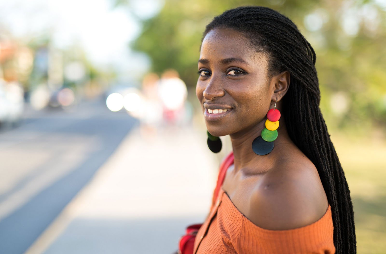 A woman with long, braided hair and colorful round earrings smiles at the camera while standing outdoors on a sunny day, wearing an off-shoulder orange top. The background is softly blurred with trees and a road.
