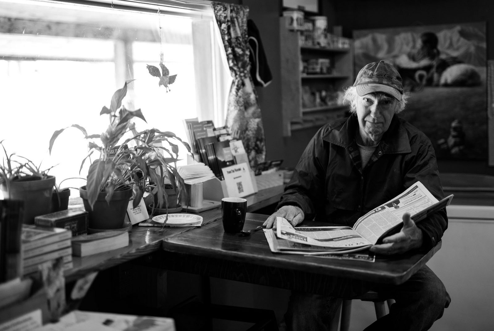An older man wearing a cap sits at a cozy wooden table by a window, reading a newspaper. Plants, books, and a coffee mug are on the table. The scene is warmly lit and filled with bookshelves and artwork in the background.