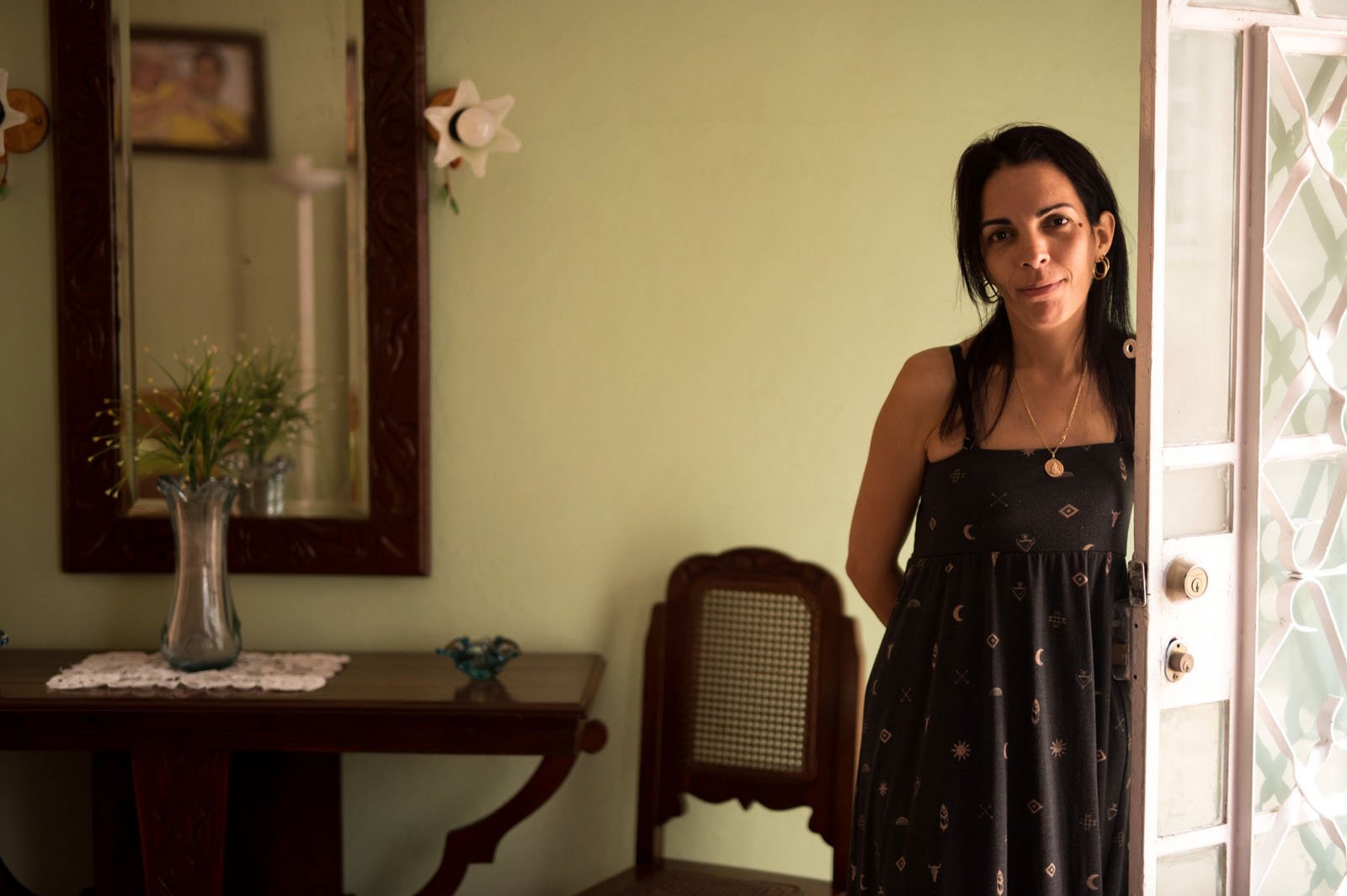 A woman in a black dress stands in a doorway, looking at the camera. Behind her is a wooden table with a vase of flowers, a chair, and a mirror hanging on a light green wall.