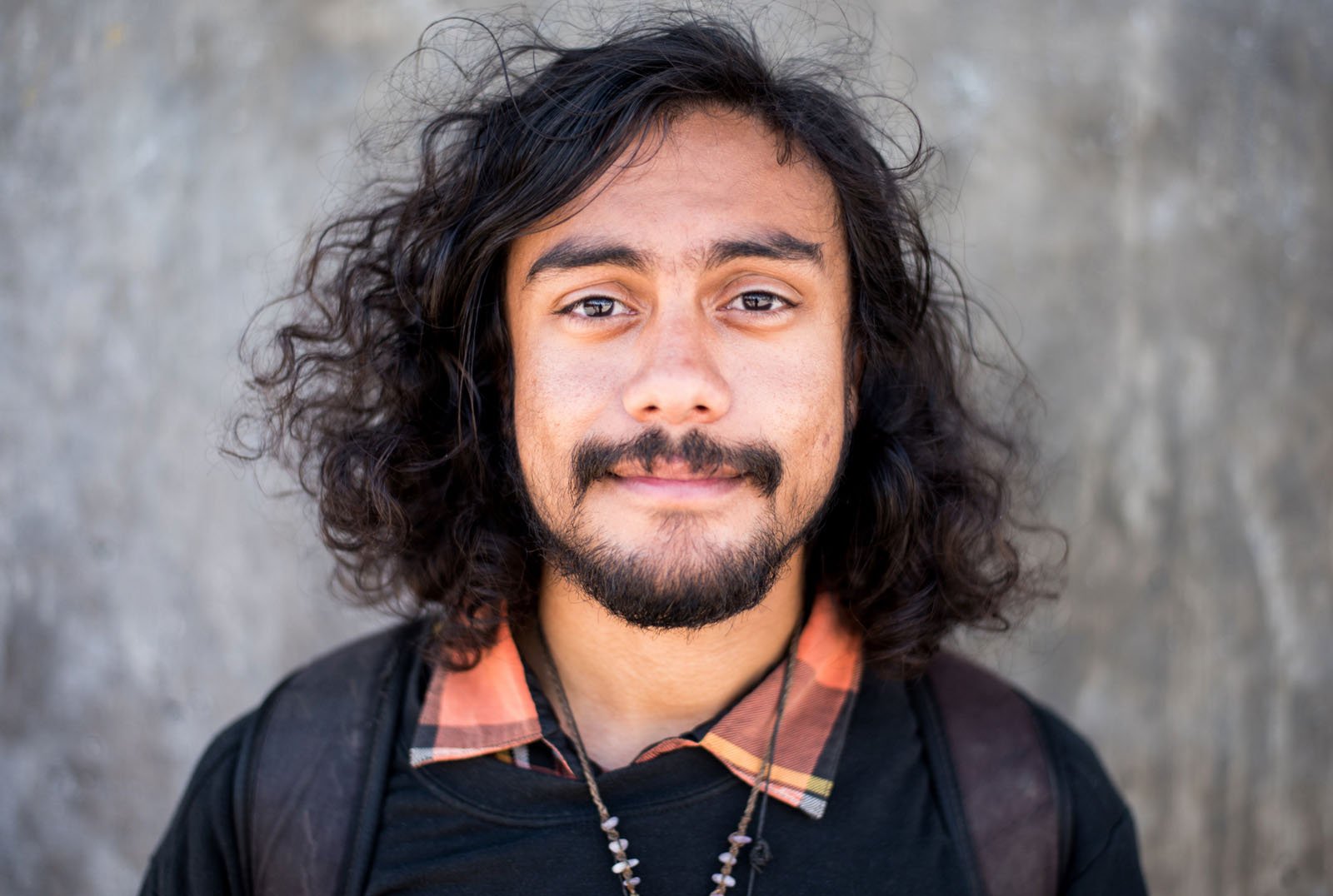 A young man with long, curly dark hair and a beard stands in front of a gray textured wall, wearing a black shirt with an orange plaid collar and a beaded necklace, looking directly at the camera with a slight smile.