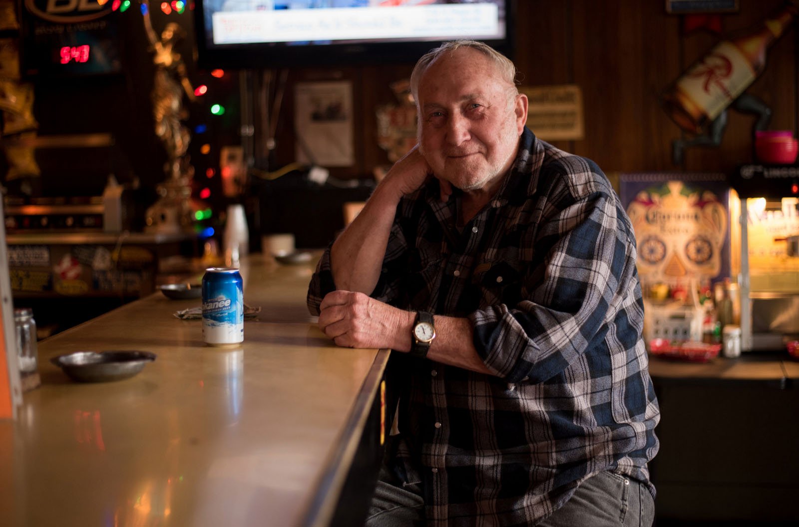 An older man in a plaid shirt sits at a dimly lit bar, smiling gently with his hand resting on his neck. A can of beer and an ashtray are on the counter in front of him. Colorful lights decorate the background.