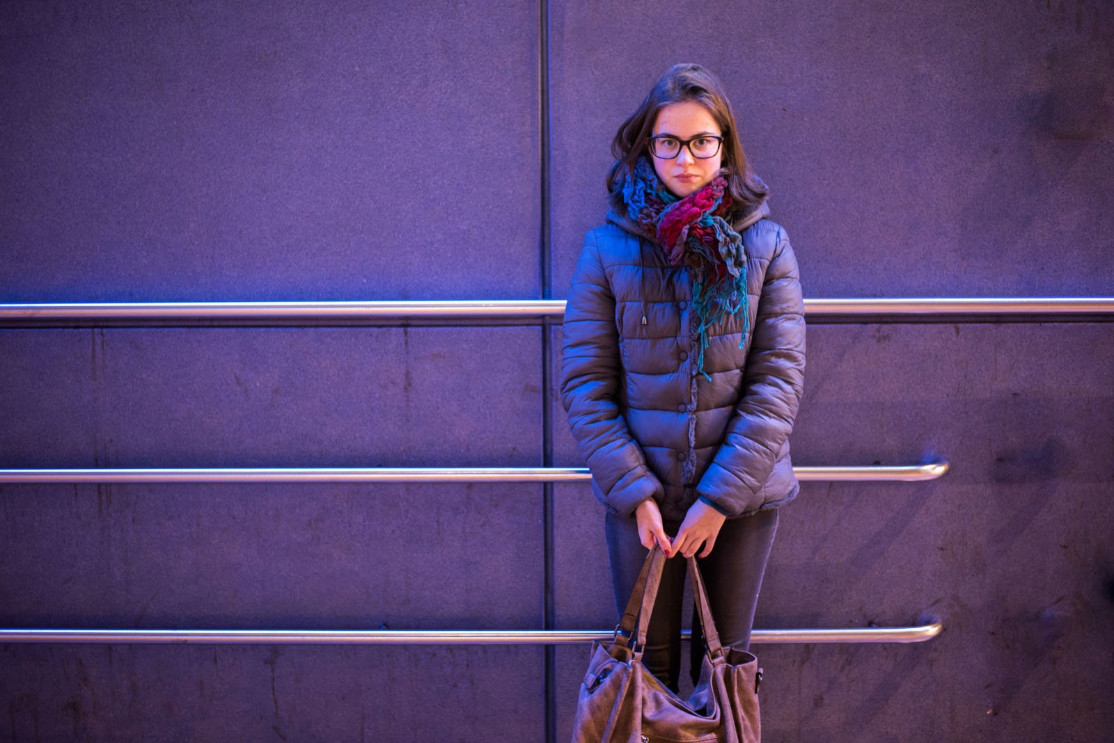 A young woman in glasses and a puffy jacket stands against a purple-lit wall, holding a brown bag with both hands and looking directly at the camera. Metal bars run horizontally along the wall behind her.