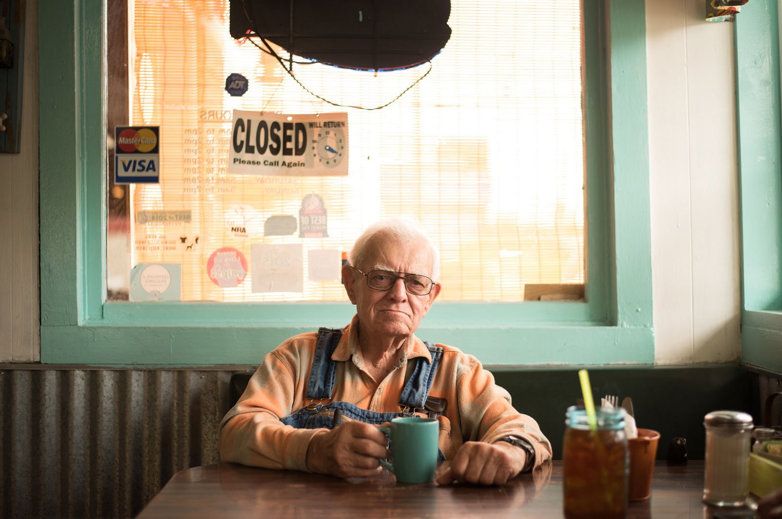 An elderly man wearing glasses and overalls sits alone at a diner table, holding a blue mug. Behind him, a window displays a "CLOSED" sign and various stickers. Sunlight filters through the blinds.