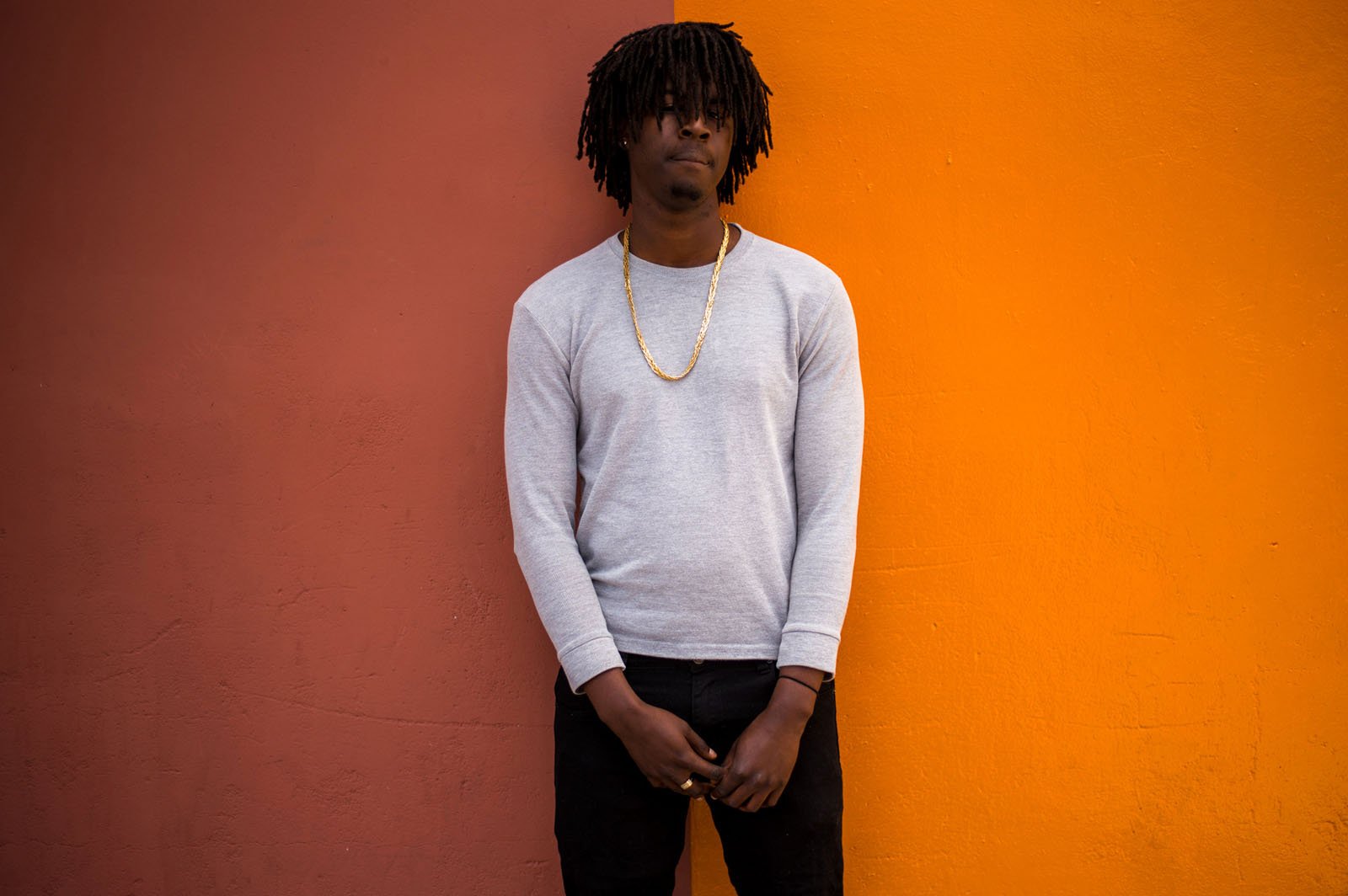 A man with medium-length dreadlocks, wearing a gray long-sleeve shirt, gold chain, and black pants, stands against a background split between reddish-brown and orange walls.