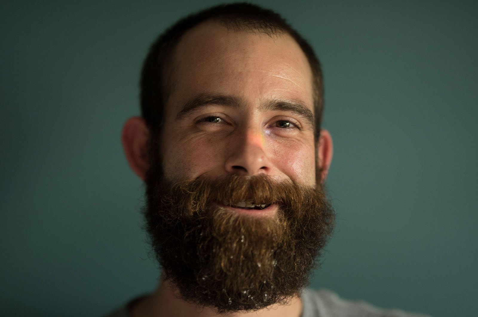 A man with a full beard and short hair smiles gently, standing against a plain teal background. The lighting is soft, highlighting his face and expression.