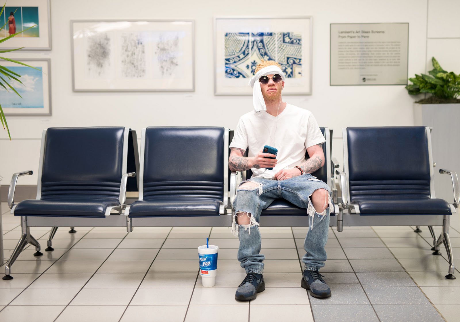 A man in casual clothes with ripped jeans and a T-shirt sits alone on blue chairs in an airport or waiting area, holding a phone. A drink cup is on the floor beside him, and art hangs on the white wall behind.
