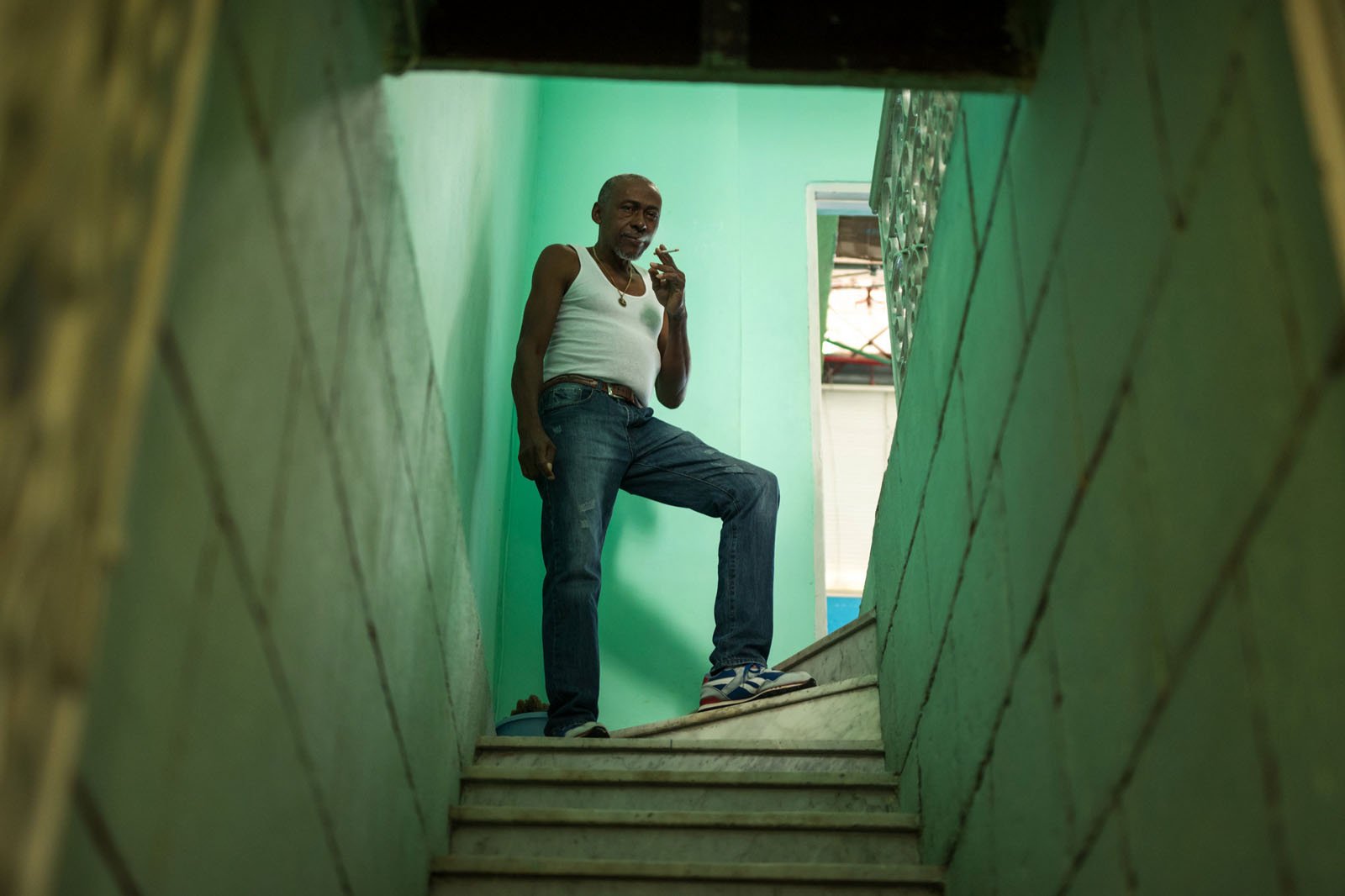 A man in a white tank top and blue jeans stands confidently at the top of a narrow stairway, smoking a cigarette, with green walls surrounding the staircase.