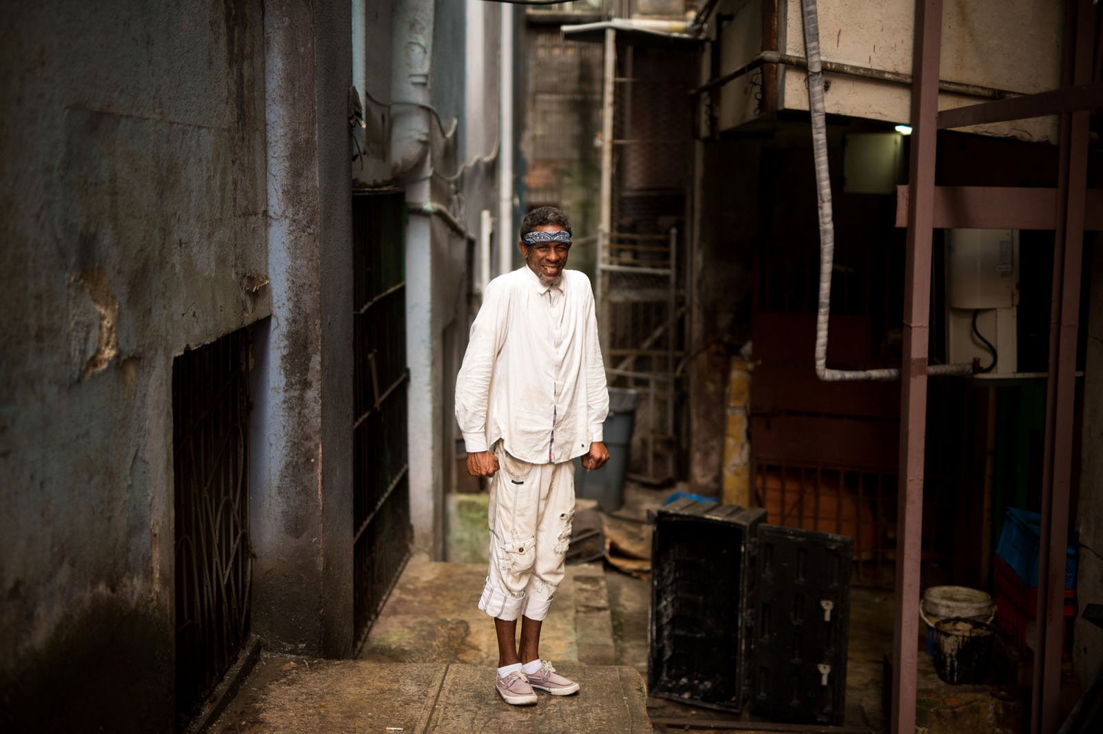 An elderly man dressed in loose, white clothing and a headband stands smiling in a narrow, urban alleyway lined with weathered walls and assorted objects.