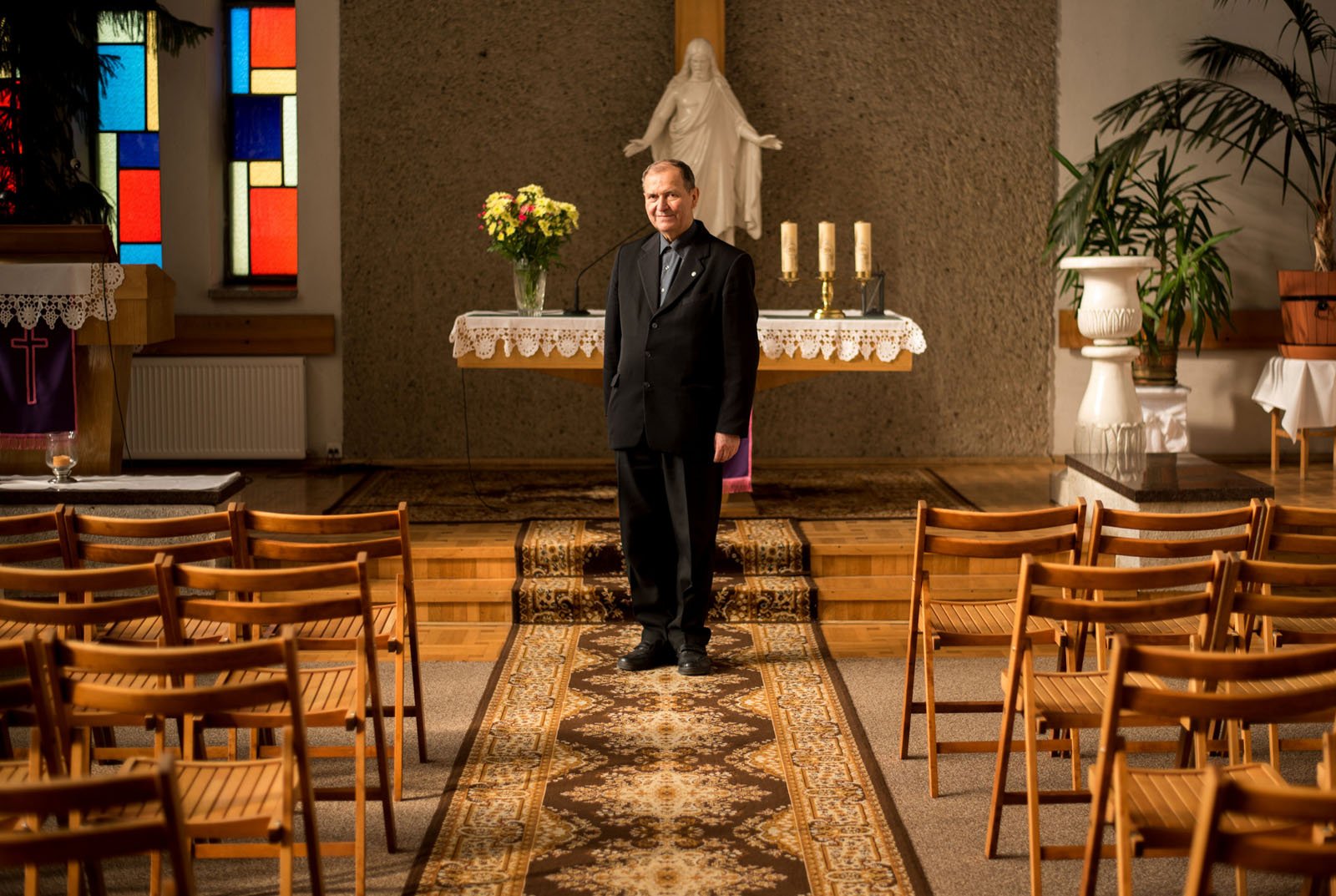 A priest in black clerical attire stands and smiles in front of an altar in a church, with empty wooden chairs arranged in rows and colorful stained glass windows visible on the left.
