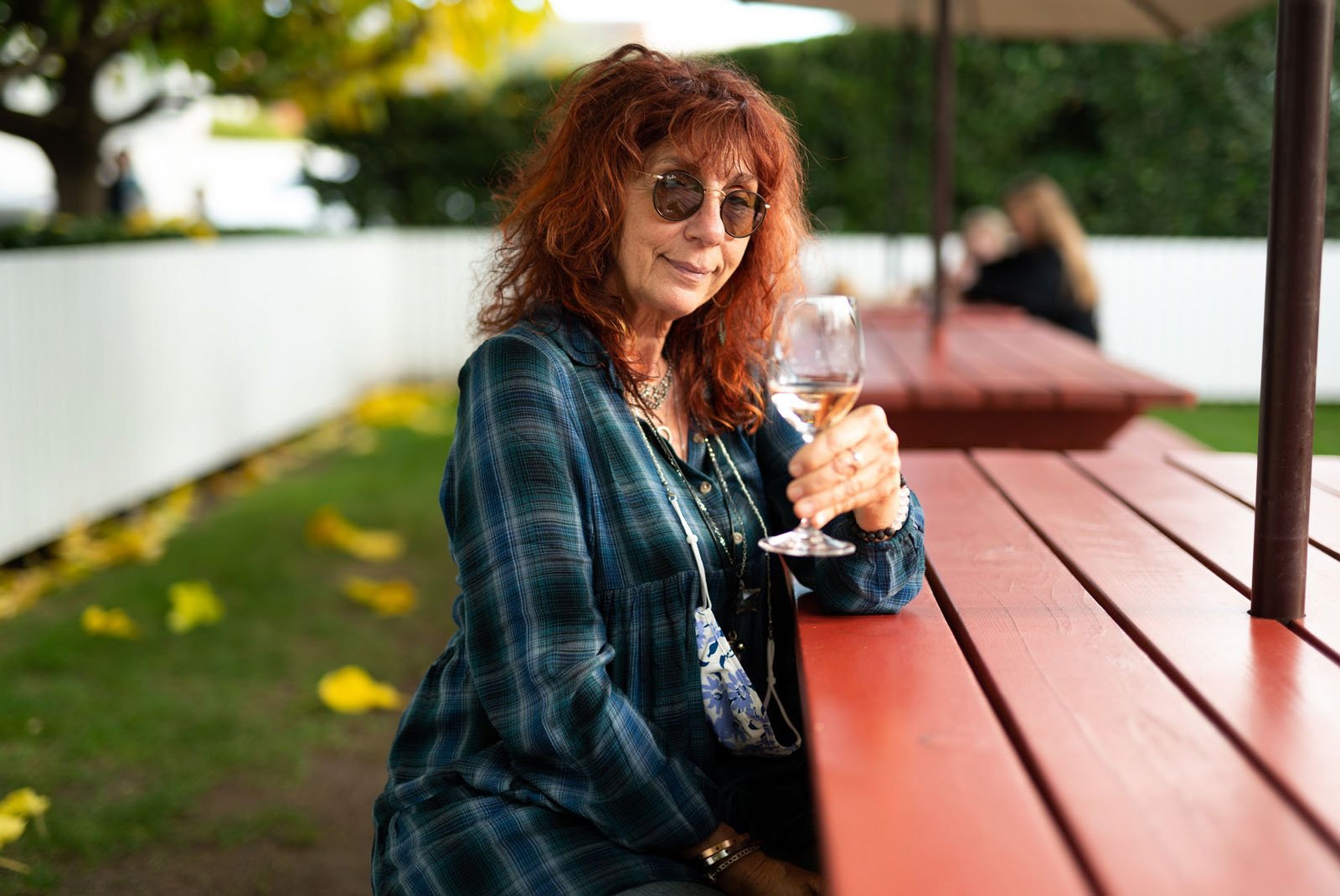 A woman with wavy red hair and sunglasses sits at a red picnic table outdoors, holding up a glass of water and smiling. Yellow leaves are scattered on the grass, and there is greenery in the background.