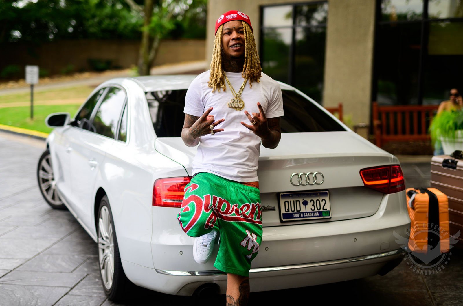 A man with long blonde braids, wearing a red cap, white t-shirt, green Supreme shorts, and sneakers, poses in front of a white Audi sedan parked on a driveway, with a suitcase and trees in the background.