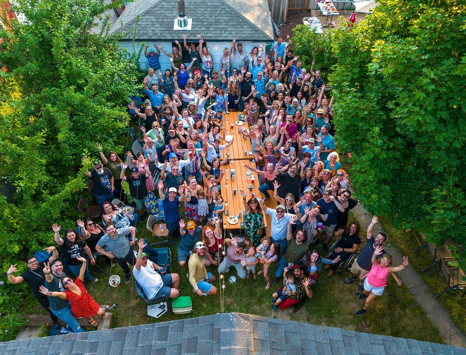A large group of people gathers around a long table in a backyard, smiling and waving up at the camera. The scene is lively, with trees surrounding the area and everyone appearing cheerful and celebratory.