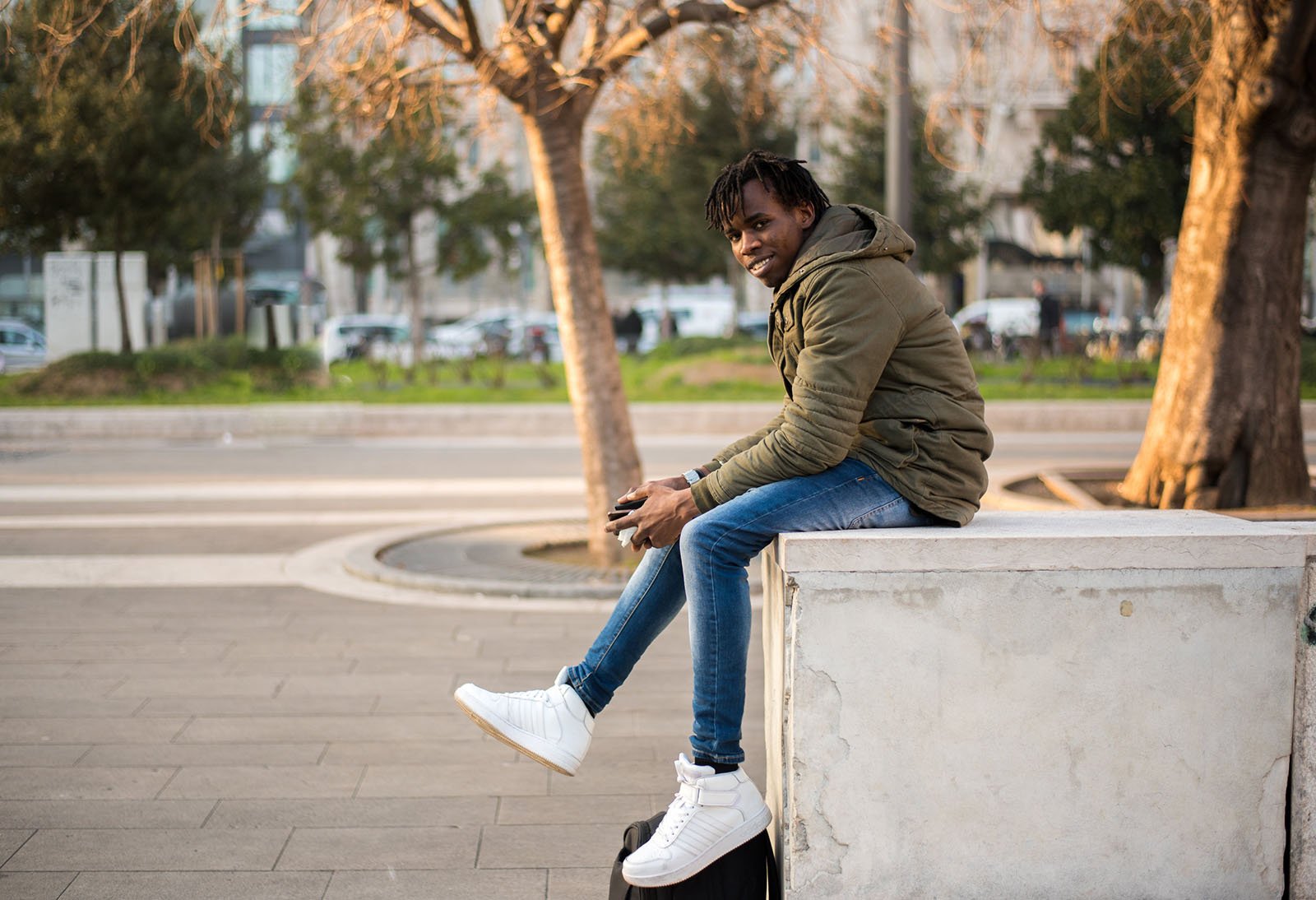A young man in a green jacket and blue jeans sits on a concrete bench outdoors, smiling at the camera. He holds a phone and wears white sneakers, with trees and parked cars in the background.