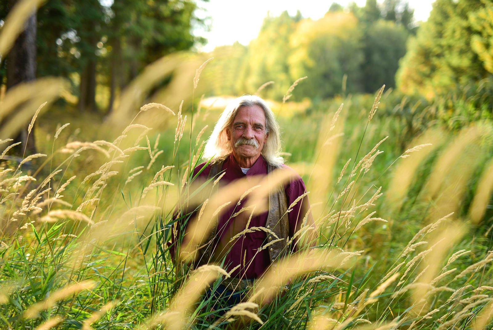 An older man with long blond hair and a mustache stands among tall grasses in a sunlit, green forest clearing, looking at the camera.