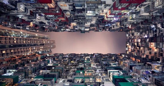 View looking up between tall, densely packed apartment buildings, creating a canyon-like effect with a narrow strip of sky visible in the center. The buildings are filled with colorful balconies and windows.