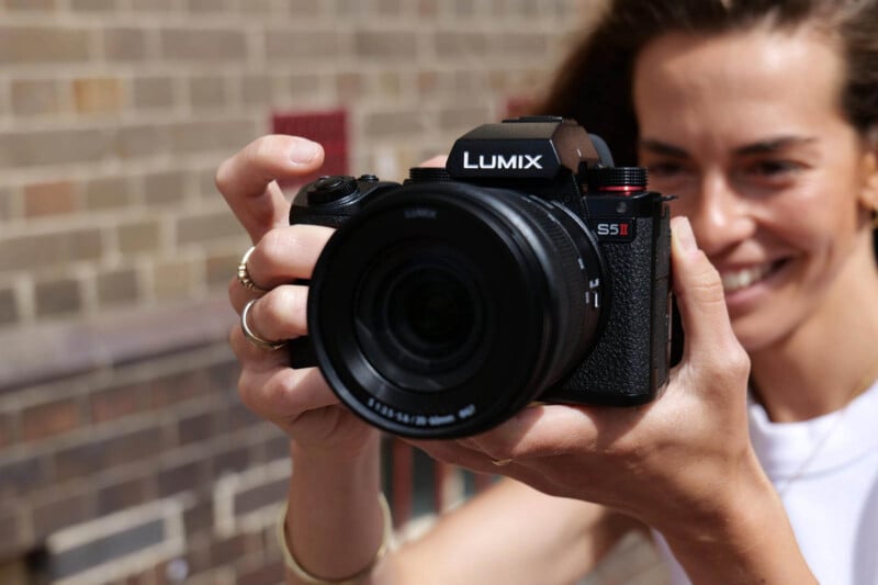 A person smiling and holding a Panasonic Lumix S5 II camera close to their face, preparing to take a photo outdoors with a blurred brick wall in the background.