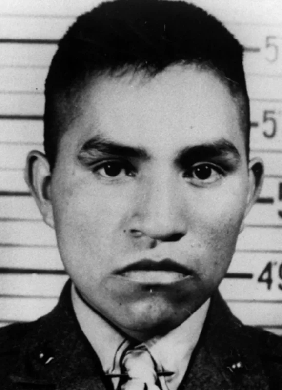 Black and white portrait of a young man in military uniform standing in front of a height chart, looking directly at the camera with a serious expression.
