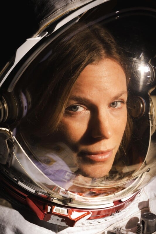 A close-up of a woman’s face inside a space helmet, with reflections on the visor and details of the spacesuit visible. She looks directly at the camera, illuminated by soft light.
