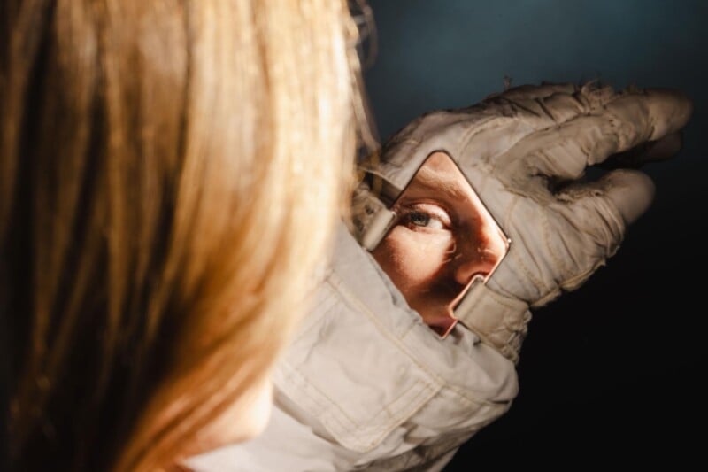 A person in an astronaut suit looks into a small rectangular mirror on their glove, with their focused eye and part of their face reflected, and their hair partially visible in the foreground.