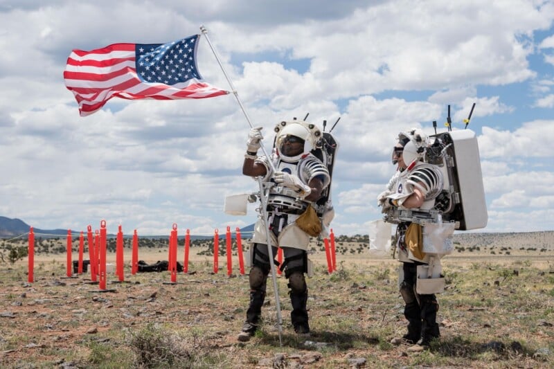 Two astronauts in spacesuits stand outdoors on rocky ground; one waves a large American flag while the other looks on. Red markers are in the background under a partly cloudy sky.