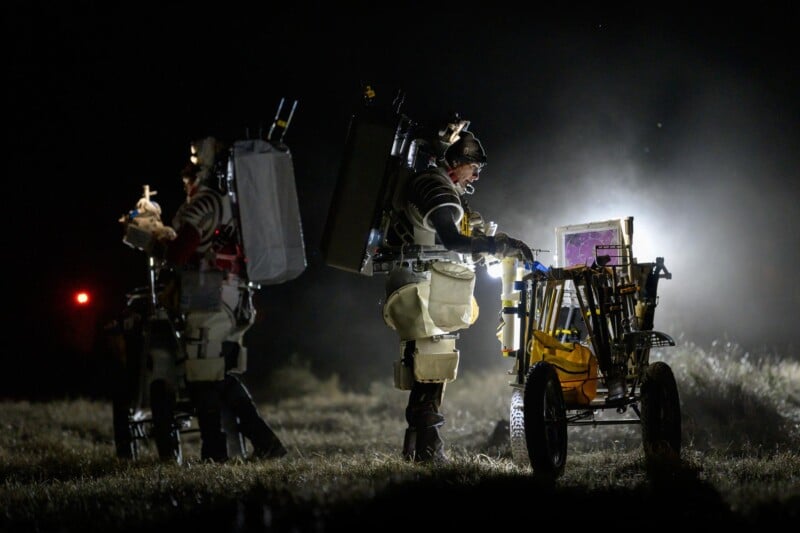 Two people in protective gear with large backpacks work at night using equipment on wheeled carts, illuminated by bright lights, in a grassy outdoor setting.