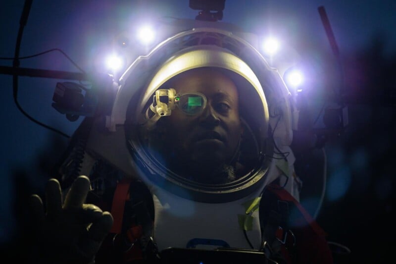 A person wearing a high-tech helmet with illuminated lights looks out from a clear visor, with their face partially obscured by reflections and shadows in a dark environment.