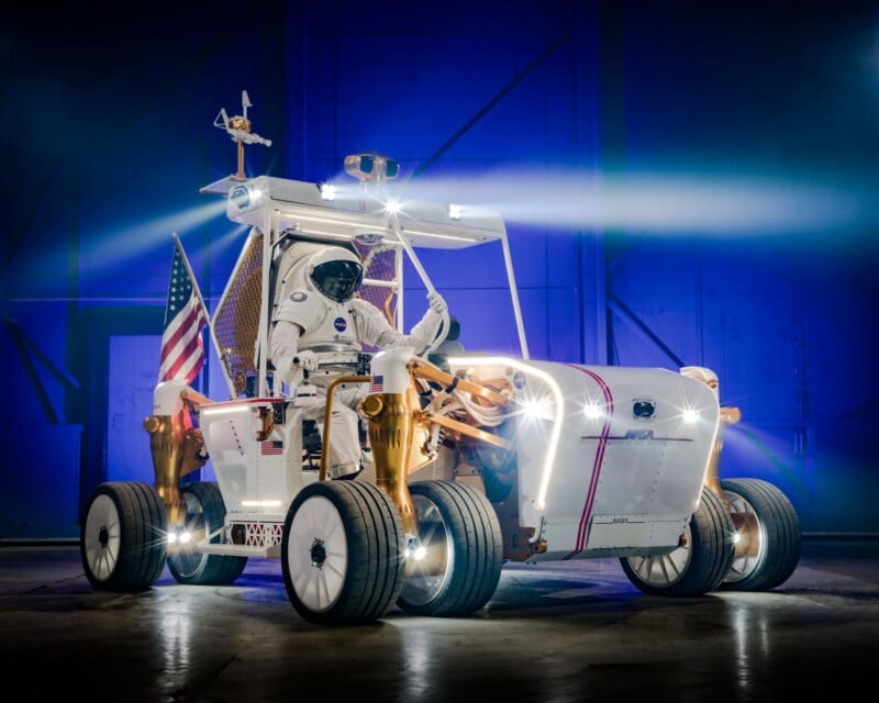 An astronaut in a spacesuit sits in a futuristic lunar rover with large wheels and bright headlights, holding an American flag, inside a dimly lit hangar with blue lighting.
