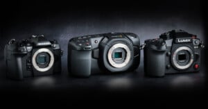 Three professional mirrorless camera bodies—Olympus, Blackmagic, and Lumix brands—are displayed side by side on a dark surface, facing forward with their lens mounts exposed.