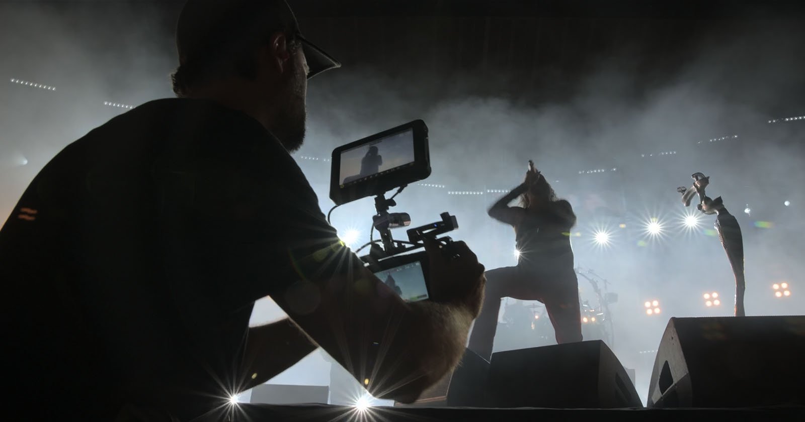 A videographer films a live concert, capturing a performer on stage under dramatic lighting and fog effects, with bright stage lights shining in the background.