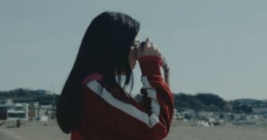 A person with long dark hair wearing a red and white jacket stands outdoors, holding a camera up to their face, with a blurred landscape and buildings in the background.