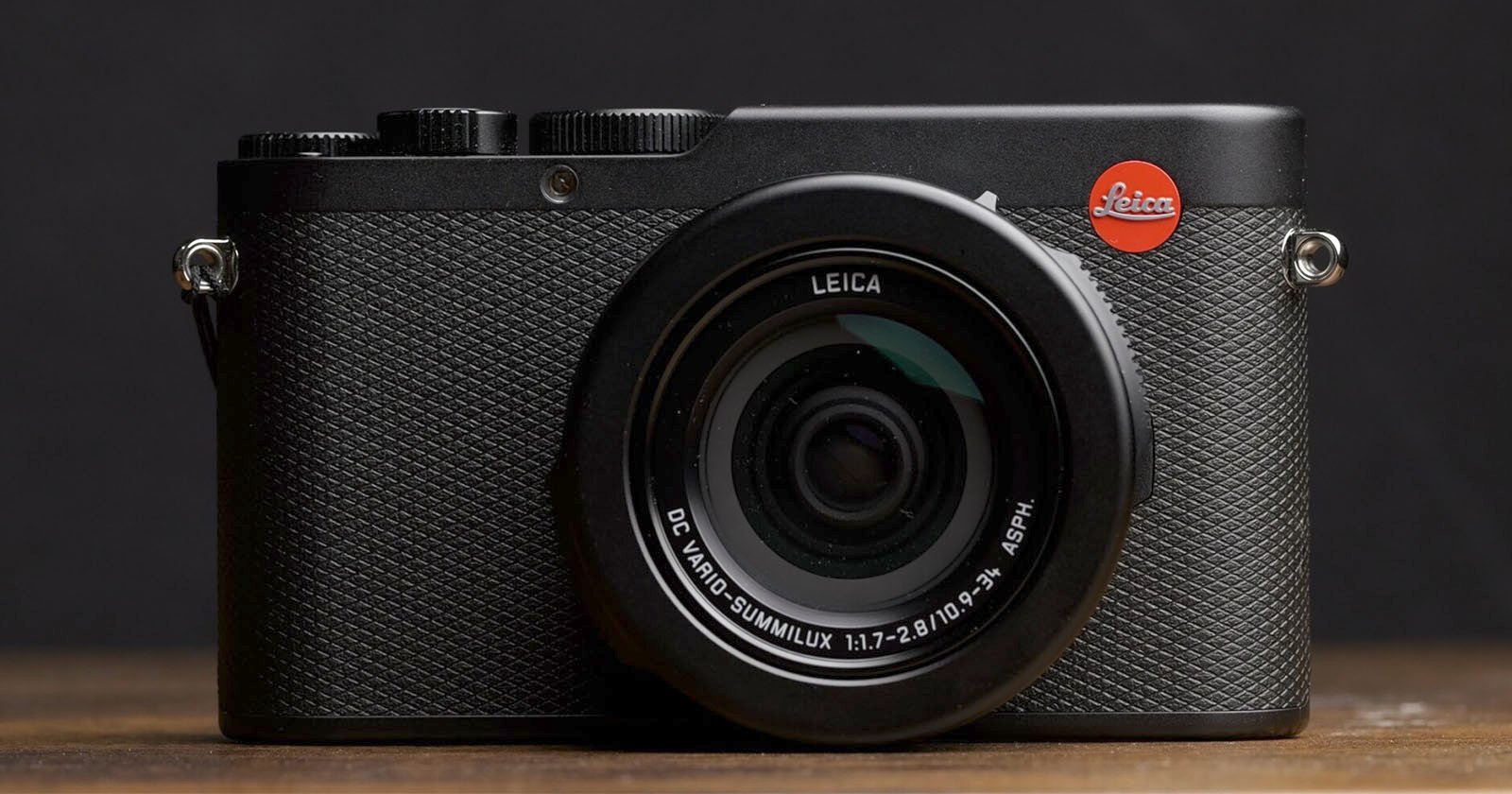 A black Leica digital camera with textured grip and red Leica logo is placed on a wooden surface against a dark background, facing forward with the lens centered.