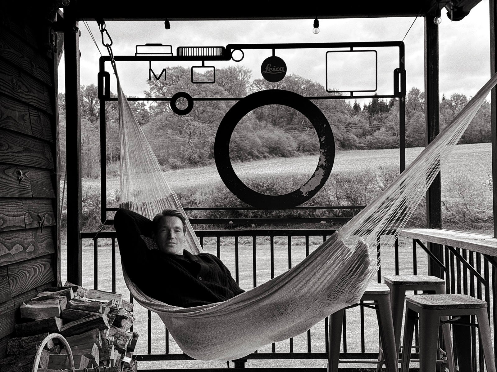 A person relaxes in a hammock on a covered porch with a large metal camera sculpture behind them, overlooking a grassy field and trees. The image is in black and white.