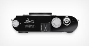 Top view of a black Leica camera, showing dials, buttons, and the lens mount; "Leica Camera New York USA" is printed on the body. The camera appears modern and sleek with a minimalist design.