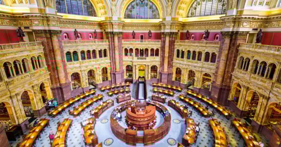A grand, ornate reading room with arched windows and tall bookshelves, featuring a circular wooden desk at the center and rows of desks with lamps arranged in a semi-circle beneath a domed ceiling.
