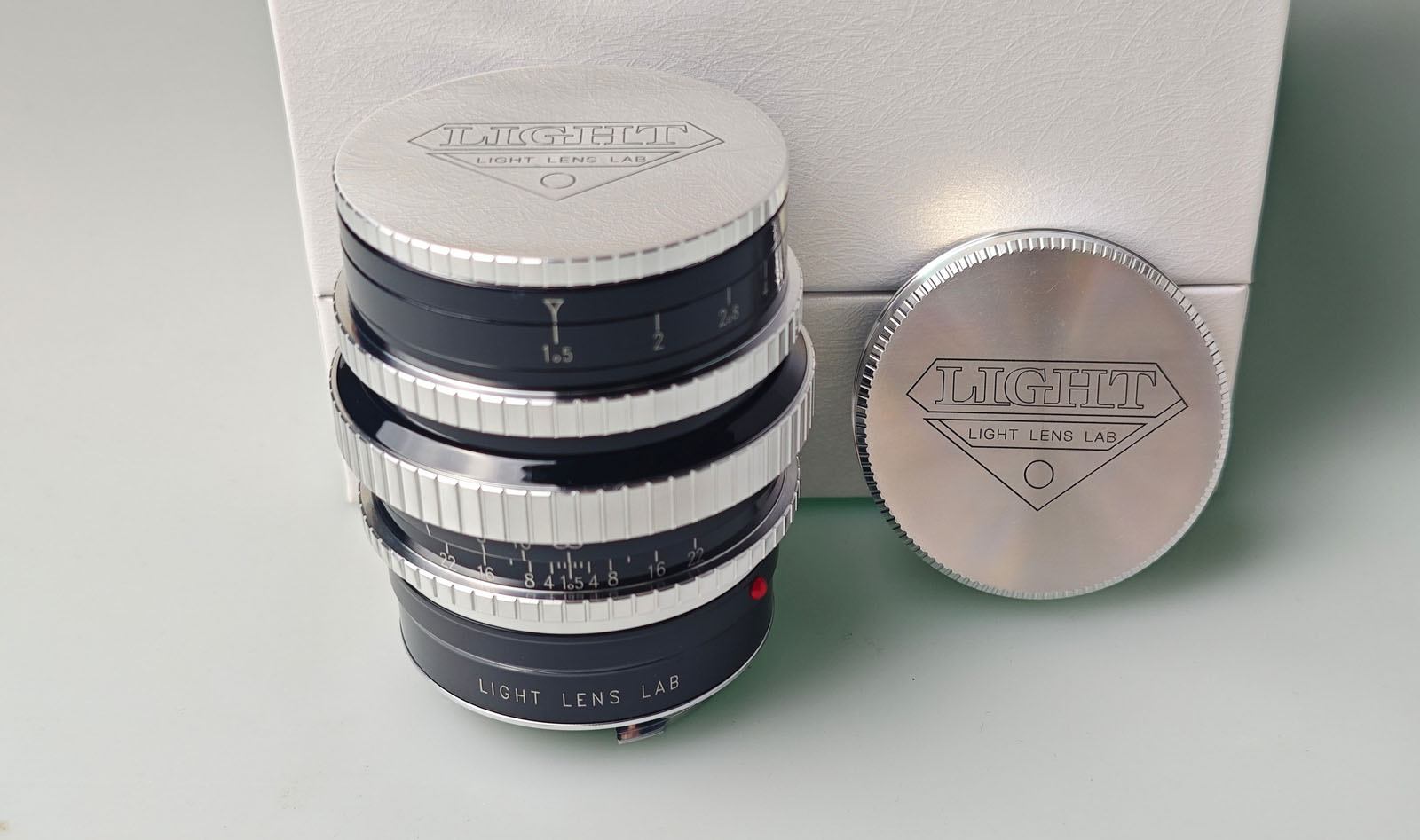 A silver and black camera lens with "LIGHT LENS LAB" branding is shown next to a matching lens cap. Both are placed on a light surface with a white background.