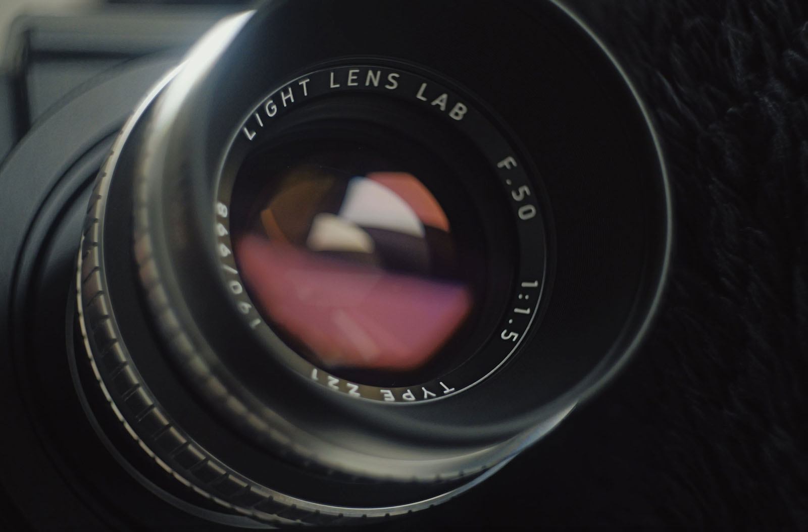 Close-up of a camera lens with text "LIGHT LENS LAB F.50 1:1.5 TYPE 271" engraved around the glass, showing reflections of light and blurred background.