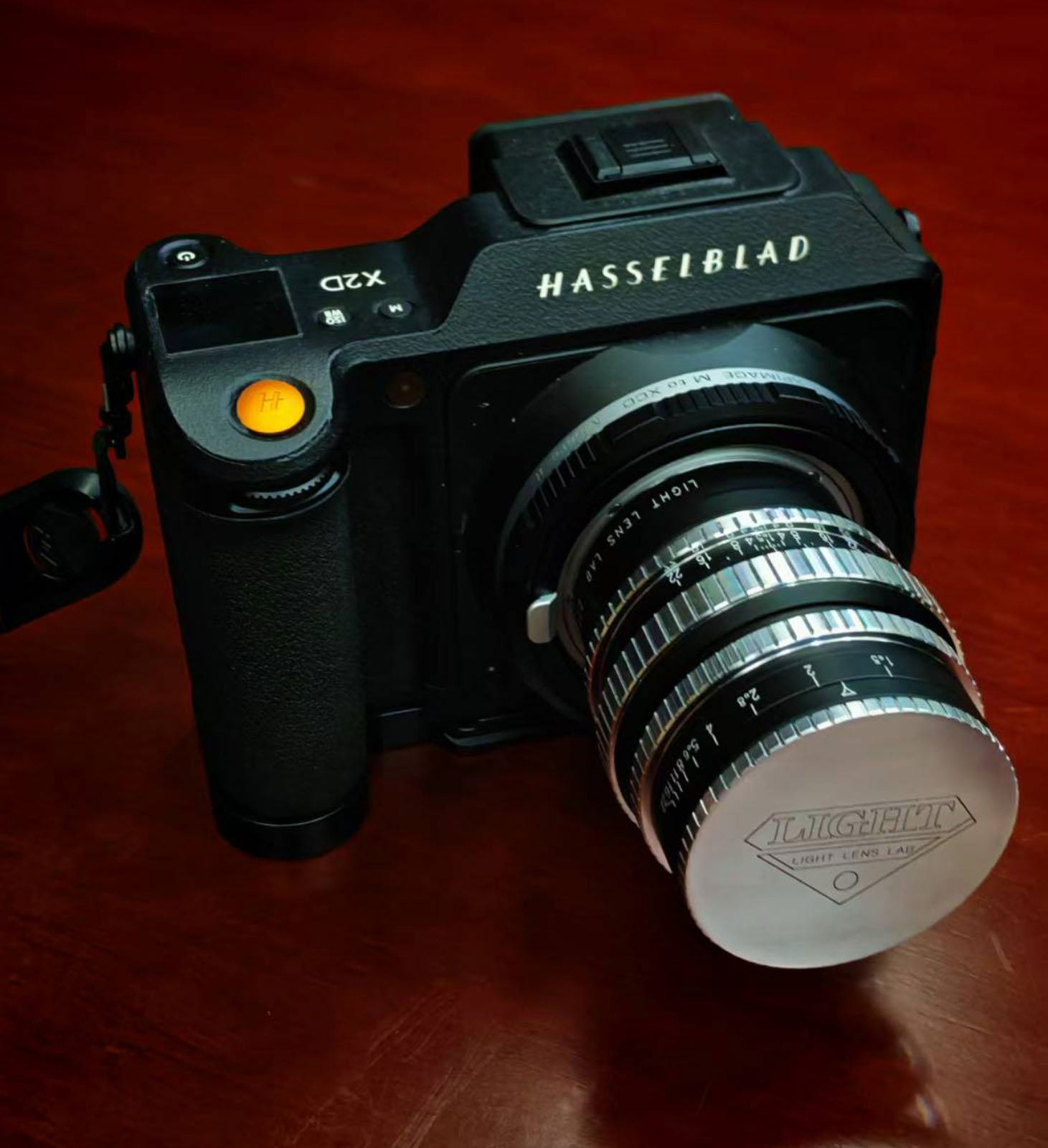 A Hasselblad X2D camera with a vintage chrome LUCHEH lens attached, resting on a reddish-brown wooden surface. The camera is angled to show the lens details and brand markings.