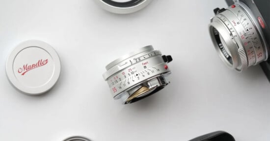 A silver camera lens with engraved numbers lies on a white surface, surrounded by lens caps and other camera equipment. The cap has "Mandle" written in red script.